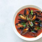 Zuppa di cozze al pomodoro facile e gustosa 1 In primo piano su uno sfondo chiaro c'รจ un piatto di zuppa di cozze con del prezzemolo fresco.