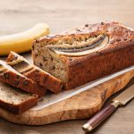 Banana bread: la ricetta facile e soffice senza burro 1 In primo piano su un tagliere in legno c'รจ un banana bread con delle gocce di cioccolato. Sullo sfondo una banana.