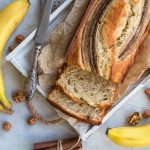 Banana Bread, la ricetta facile e soffice del dolce alla banana 1 su tavolo con tovaglia, primo piano di una teglia con il banana bread con 2 fette tagliate e sul bordo un coltello. A destra, 2 banane intere e a sinistra 1. Attorno frutta secca sparsa