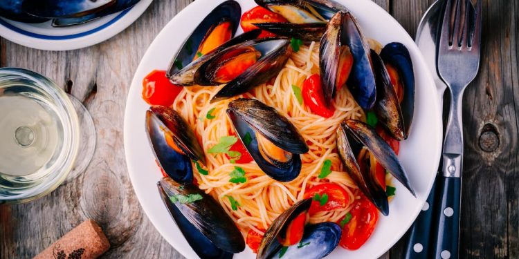 su un piano di cucina primo piano di un piatto di spaghetti con le cozze, un bicchiere di vino, una forchetta e un coltello e un tappo di bottiglia