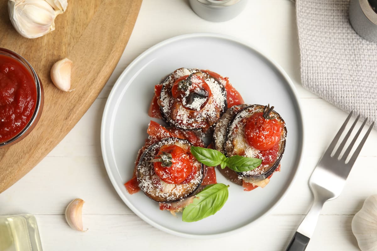 In primo piano su un piatto fondo bianco ci sono delle porzioni di melanzane alla parmigiana decorate con delle foglie di basilico.