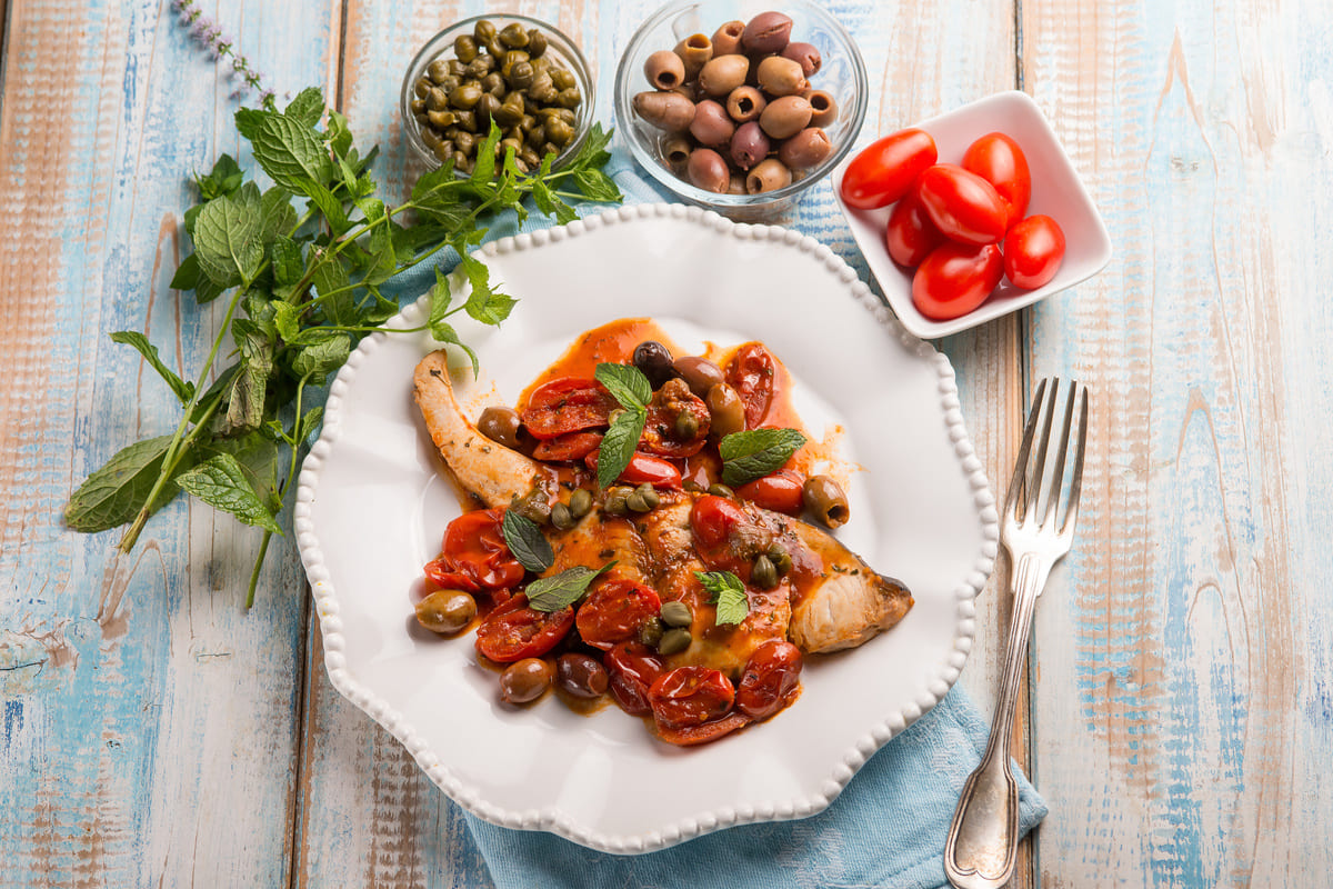 Su un tavolo di legno c'è un piatto bianco con del pesce spada alla siciliana pomodorini, olive e capperi. Intorno al piatto c'è un rametto di basilico, dei capperi, olive e pomodorini.