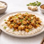 Pollo alle mandorle: secondo piatto della cucina cinese in padella 1 In primo piano su un tavolo chiaro c'è un piatto con del pollo alle mandorle e sulla base del riso condito con dei porri. Sullo sfondo, a sinistra in una ciotola c'è del riso mentre a destra c i sono delle mandorle e una radice di zenzero.