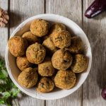 Polpette di melanzane al forno, morbide e saporite 1 In primo piano su un tavolo di legno c'รจ un piatto con delle polpette di melanzane. Sullo sfondo ci sono delle melanzane fresche, una testa d'aglio e del prezzemolo.