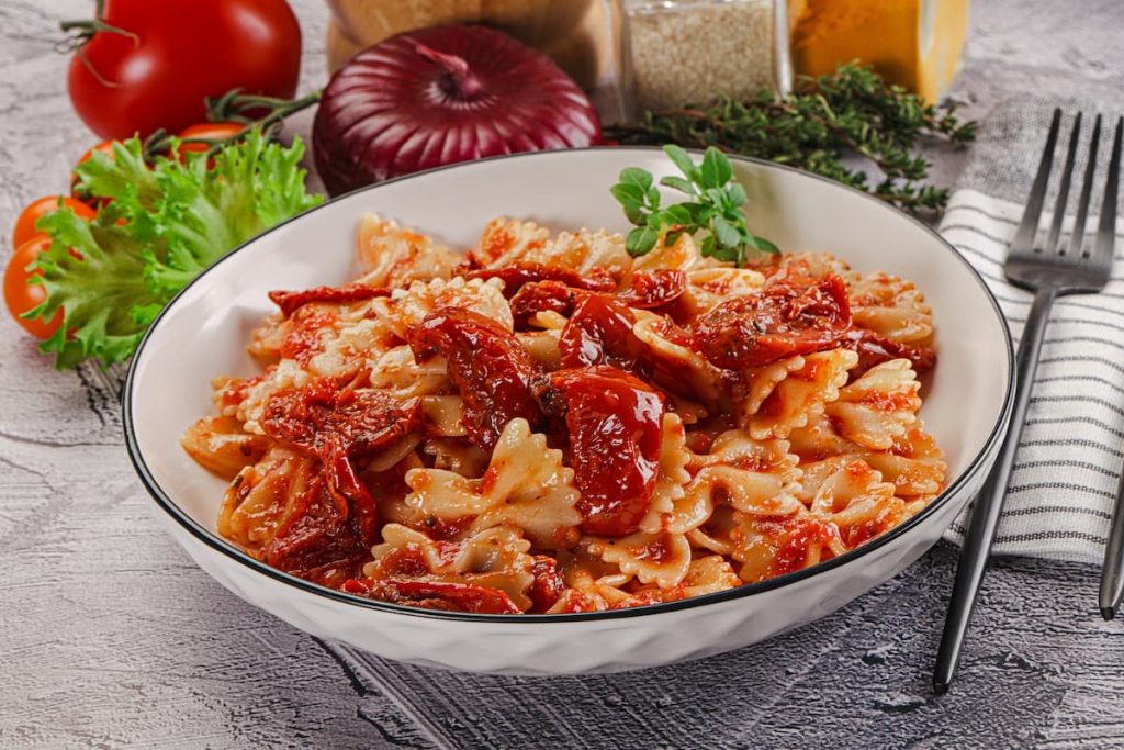 piano di cucina con piatto fondo con pasta con pomodori secchi, tovagliolo piegato e una forchetta, e ingredienti della preparazione
