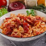 piano di cucina con piatto fondo con pasta con pomodori secchi, tovagliolo piegato e una forchetta, e ingredienti della preparazione