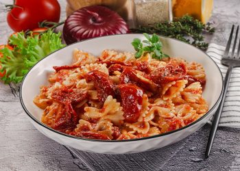 piano di cucina con piatto fondo con pasta con pomodori secchi, tovagliolo piegato e una forchetta, e ingredienti della preparazione
