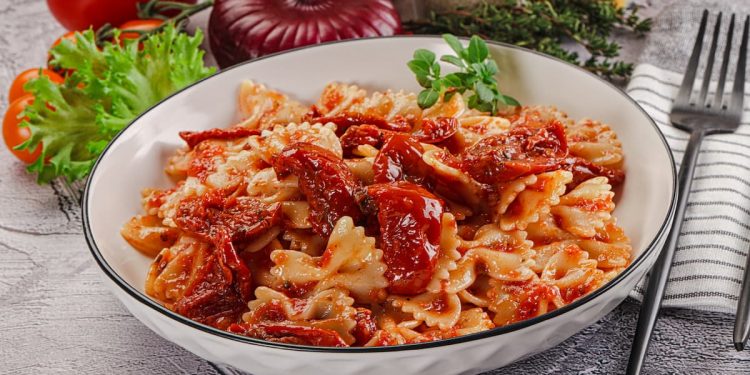 piano di cucina con piatto fondo con pasta con pomodori secchi, tovagliolo piegato e una forchetta, e ingredienti della preparazione