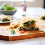 su tavolo di legno bianco un tagliere con la Spanakopita tagliata e fuori fuoco un piatto di spinaci freschi