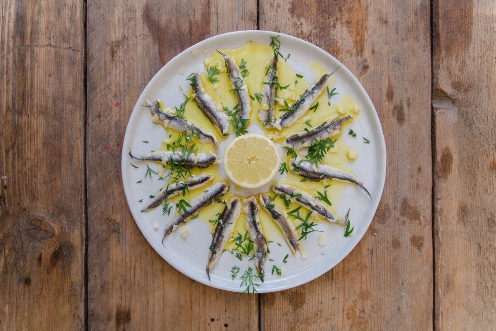 su tavolo di legno, piatto bianco con alici marinate, olio Evo e una fetta di limone al centro