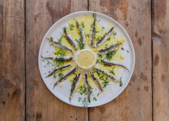 su tavolo di legno, piatto bianco con alici marinate, olio Evo e una fetta di limone al centro