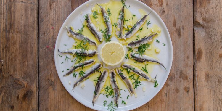 su tavolo di legno, piatto bianco con alici marinate, olio Evo e una fetta di limone al centro