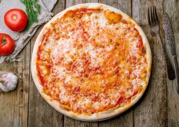 su un tavolo di legno una pizza margherita con forchetta e coltello, aglio e pomodori per far capire le calorie della pizza