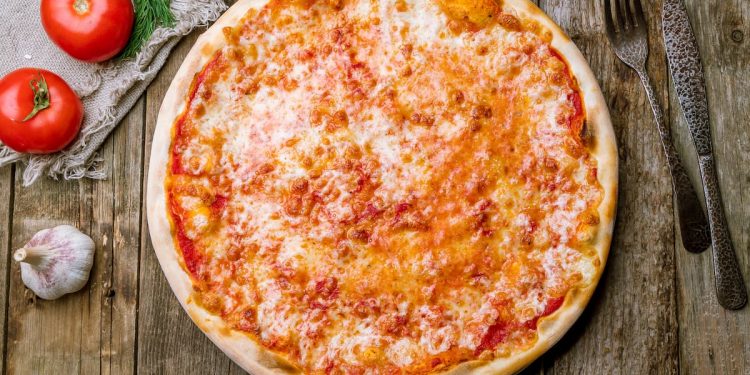 su un tavolo di legno una pizza margherita con forchetta e coltello, aglio e pomodori per far capire le calorie della pizza