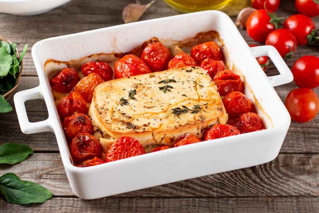 Feta al forno: un piatto vegetariano greco 2 su tavolo di legno, piatto da forno bianco con feta al forno e pomodorini