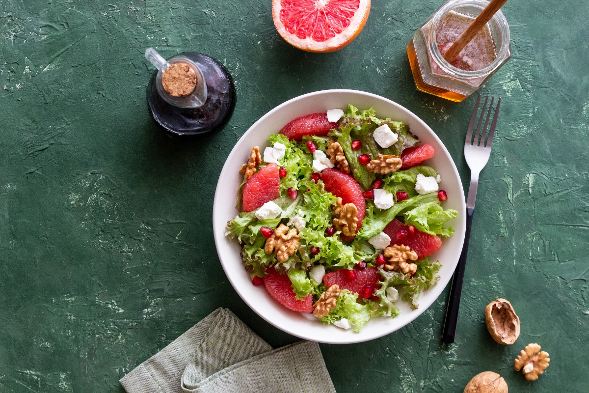 Su tavolo verde, ciotola con insalata verde, pomplemo rosa, noci e uno dei formaggi magri come caprino, attorno i condimenti