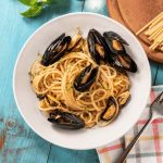 Spaghetti con le cozze: ricetta cremosa e saporita 1 In primo piano, su un tavolo di legno azzurro, c’è un piatto di spaghetti con le cozze. Sulla destra si vede un tovagliolo a quadri colorati, mentre sullo sfondo ci sono spaghetti e prezzemolo fresco.