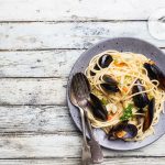 su un tavolo di legno bianco un piatto grigio con gli spaghetti con le cozze una forchetta e un cucchiaio