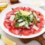 Carpaccio di manzo: ricetta fresca e delicata 1 In primo piano su un piatto fondo chiaro c'è un carpaccio di manzo condito con della rucola, dei capperi e scaglie di parmigiano.
