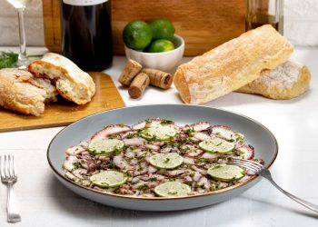 su tavolo con tovaglia bianca, piatto grigio con un carpaccio di polpo e zucchine e dietro pane, un bicchiere di vino e limoni