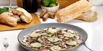 su tavolo con tovaglia bianca, piatto grigio con un carpaccio di polpo e zucchine e dietro pane, un bicchiere di vino e limoni