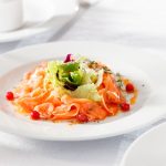 In primo piano su un tavolo bianco c'è un piatto fondo con del carpaccio di salmone e dell'insalata fresca al centro.