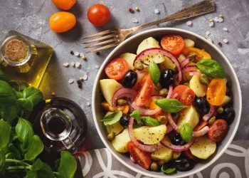 su tavolo una ciotola con l'insalata pantesca con patate pomodorini, capperi e cipolle rosse, accanto olio EVO e basilico
