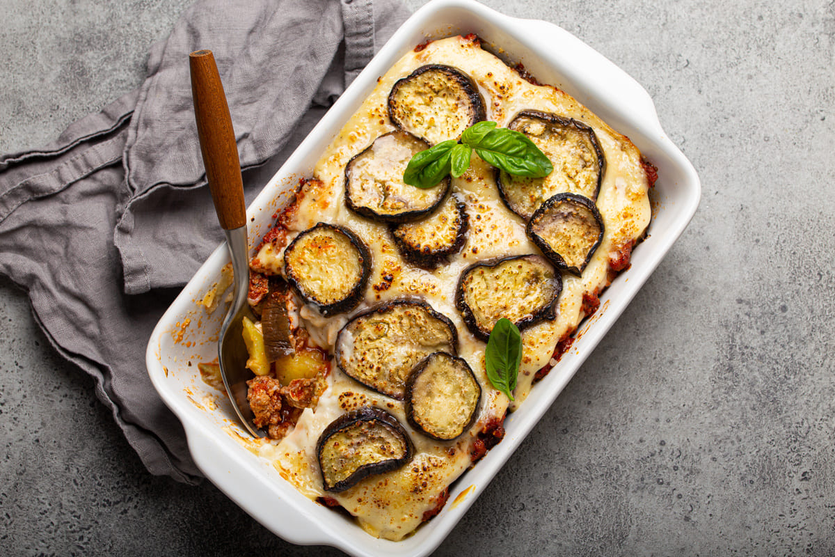 In primo piano su uno sfondo grigio c'è una pirofila bianca con la moussaka di melanzane.