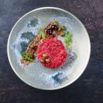 Tartare di manzo: ricetta facile e pronta in 10 minuti 1 In primo piano su uno sfondo scuro, c'è un piatto chiaro con la tartare di manzo, dei capperi e delle olive nere.