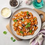 Pasta alla Norma: la ricetta originale facile e gustosa 1 Pasta alla Norma con penne, melanzane, ricotta salata e basilico in un piatto su tagliere, con ciotole di pepe e formaggio sullo sfondo.