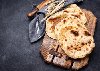 su sfondo nero, tagliere di legno con 4 pani pita, un mattarello