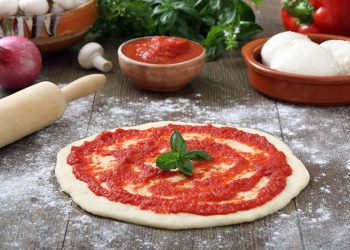 su piano da cucina di legno, pizza rossa cruda con passata di pomodori, un mattarello, cipolla, basilico, una ciotola con 2 mozzarelle