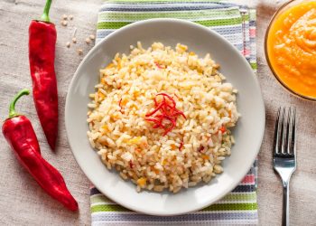 su tavolo con tovaglia beige piatto bianco con riso parboiled, 2 peperonicini, una ciotola con purée di carote e una forchetta
