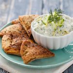 Tzatziki: la ricetta originale greca fresca e leggera 1 su tavolo di legno, piatto azzurro con una ciotola di salsa tzatziki con limone e aneto e attorno pane pita
