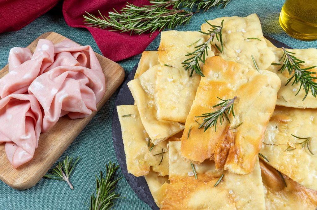 Scrocchiarella: la pizza romana croccante e deliziosa 3 vassoio con pizza bianca scrocchiarella e rosmarino, vassoio con mortadella accanto
