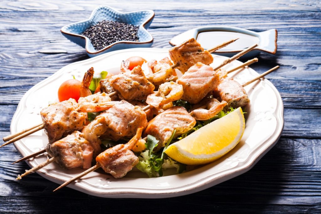 Spiedini di pesce: la ricetta e come cucinarli 3 su tavolo di legno piatto bianco con spiedini di pesce e una fetta di limone, in secondo piano pepe nero
