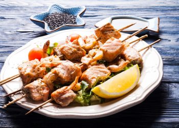 su tavolo di legno piatto bianco con spiedini di pesce e una fetta di limone, in secondo piano pepe nero