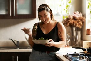 Iscriviti subito alla dieta Melarossa e scarica l'app 15 una donna piena di tatoo è sorridente e è appoggiata sul banco della cucina e ha nel suo piatto un'insalata perché sto seguendo una dieta per trigliceridi alti