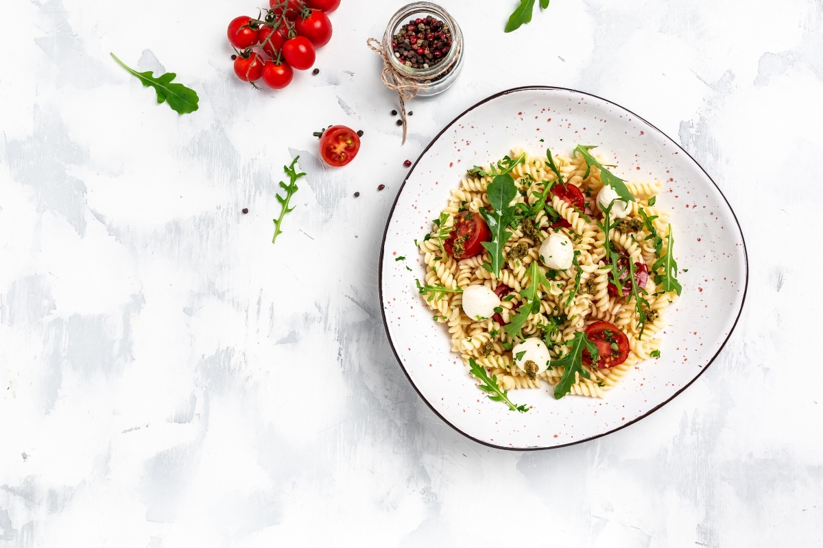 su piano da cucina di marmo bianco un piatto bianco con pasta fredda con pomodorini, rucola e bocconcini di mozzarella. Dietro un contenitore di pepe e un grappolo di pomodorini