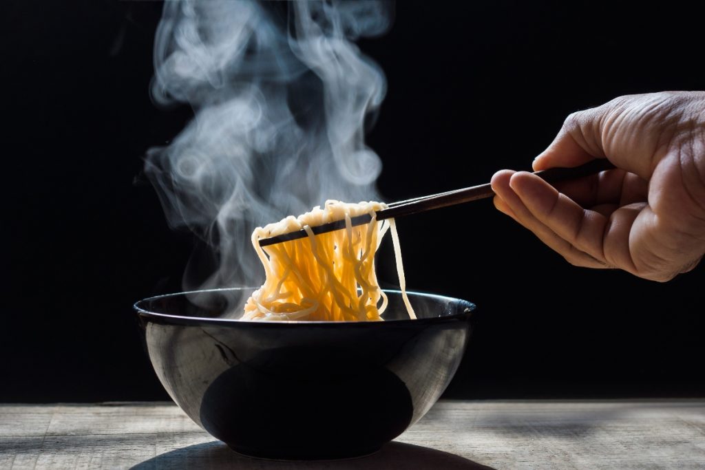Noodles fatti in casa: la ricetta degli spaghetti giapponesi 5 su tavolo ciotola nera con noodles fatti in casa che fumano, una mano li prende con le bacchette cinesi