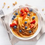 su base bianca una scodella con 4 pancake proteici con sopra fragole, mirtilli e frutta secca