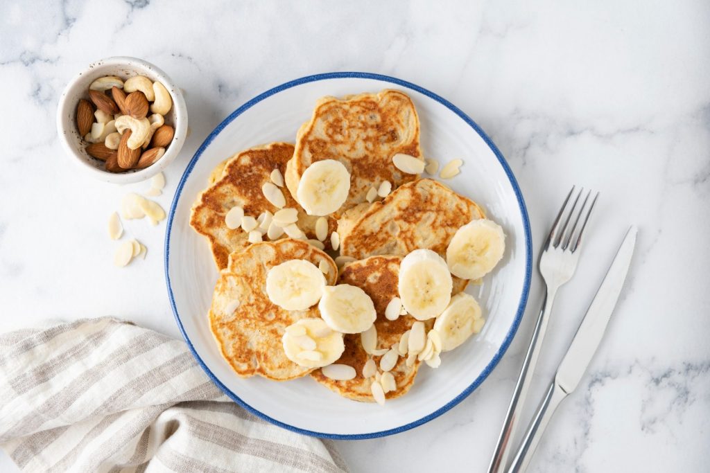 Pancake proteici: la ricetta gustosa per una colazione ricca di proteine 3 su piano di marmo, piatto con pancake proteici e banane tagliata, accanto forchetta e coltello e frutta secca