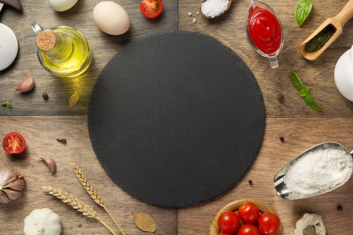 In primo piano su un tavolo in legno c'è una pietra refrattaria, intorno ci sono gli ingredienti principali per condire una pizza tra cui olio evo, farina, pomodorini e basilico.