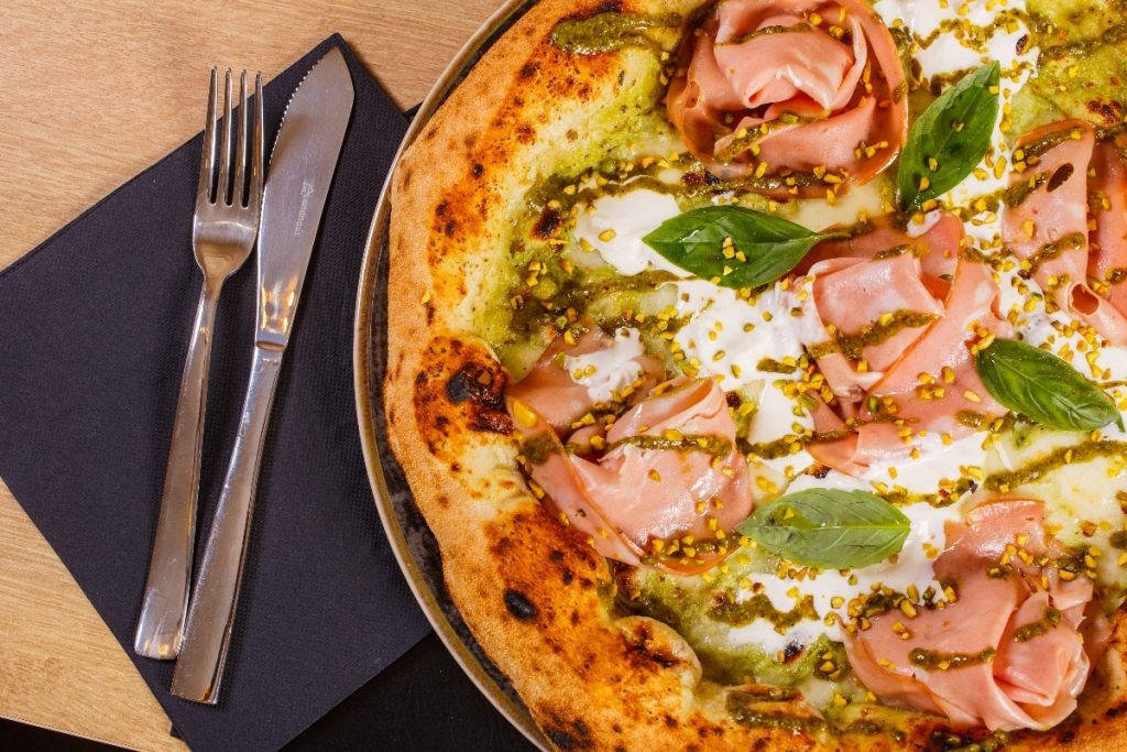 Pizza mortadella e pistacchio: ricetta e segreti di una pizza gourmet 1 primo piano di una pizza mortadella e pistacchi con mozzarella su un tavolo e accanto tovagliolo di carta blu con forchetta e coltello