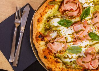 primo piano di una pizza mortadella e pistacchi con mozzarella su un tavolo e accanto tovagliolo di carta blu con forchetta e coltello