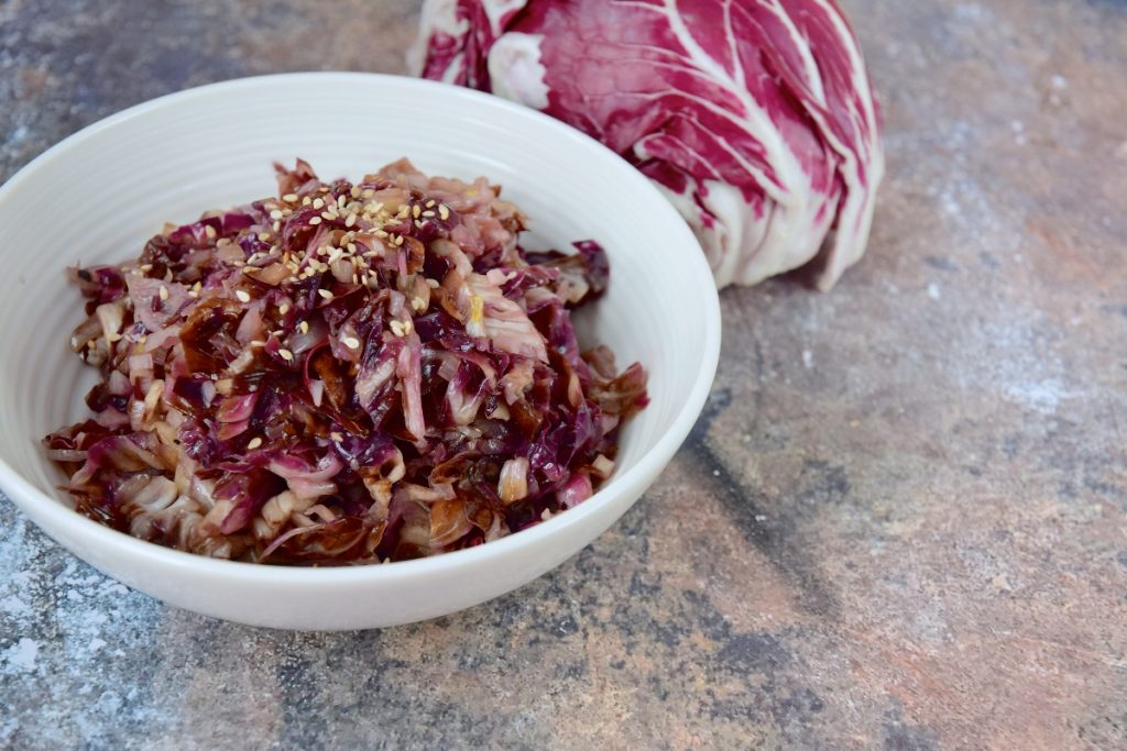 Radicchio in padella: la ricetta gustosa per un contorno perfetto 2 su tavolo di marmo, radicchio in padella in una ciotola bianca con semi di sesamo e dietro un radicchio