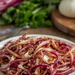 Radicchio in padella: la ricetta e video ricetta di un contorno veloce 1 In primo piano su un tavolo di legno un piatto bianco con del radicchio in padella. Sullo sfondo delle posate, radicchio e aglio.