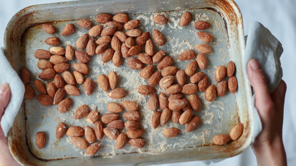 Mandorle tostate: uno snack sano e versatile 3 primo piano di mani che tengono una teglia da forno con carta da forno e mandorle tostate