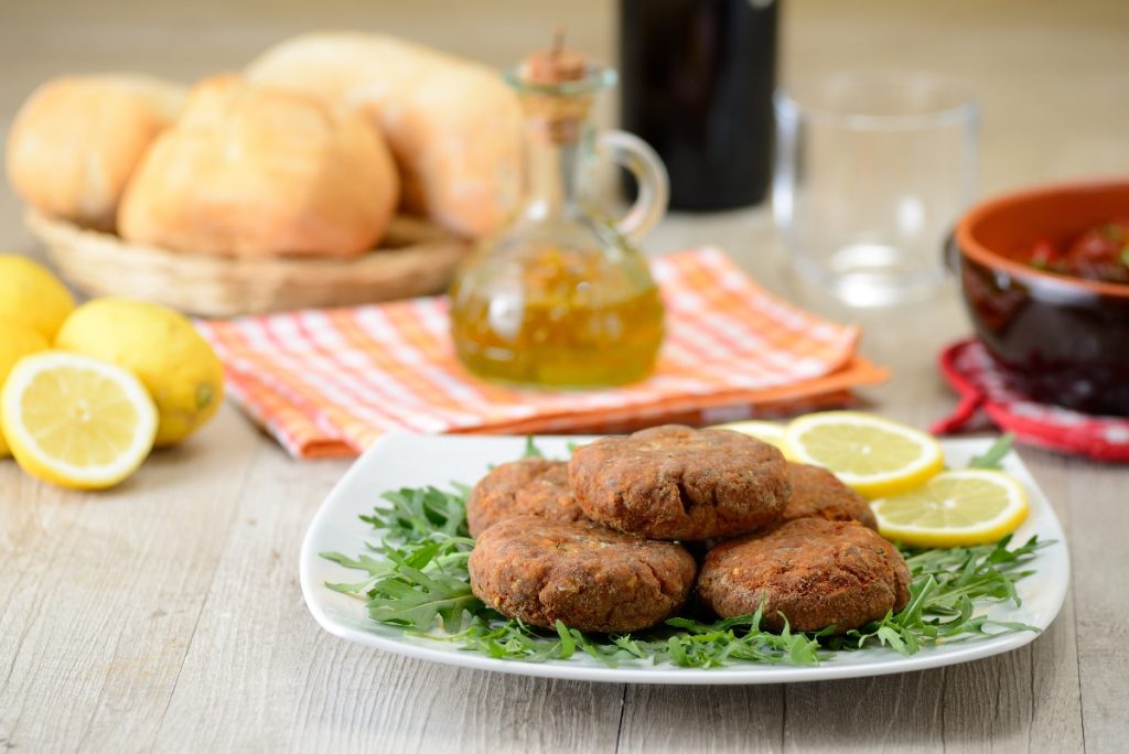 Mondeghili: la ricetta delle tipiche polpette milanesi 4 su tavolo da cucina, piatto con rucola, limoni tagliati e mondeghili, dietro una bottiglietta di olio EVO e panini