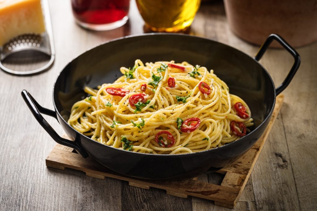 Su tavolo di legno, tagliere, una padella con Spaghetti aglio, olio e peperoncino , dietro una grattugia e un bicchiere di olio d'olive